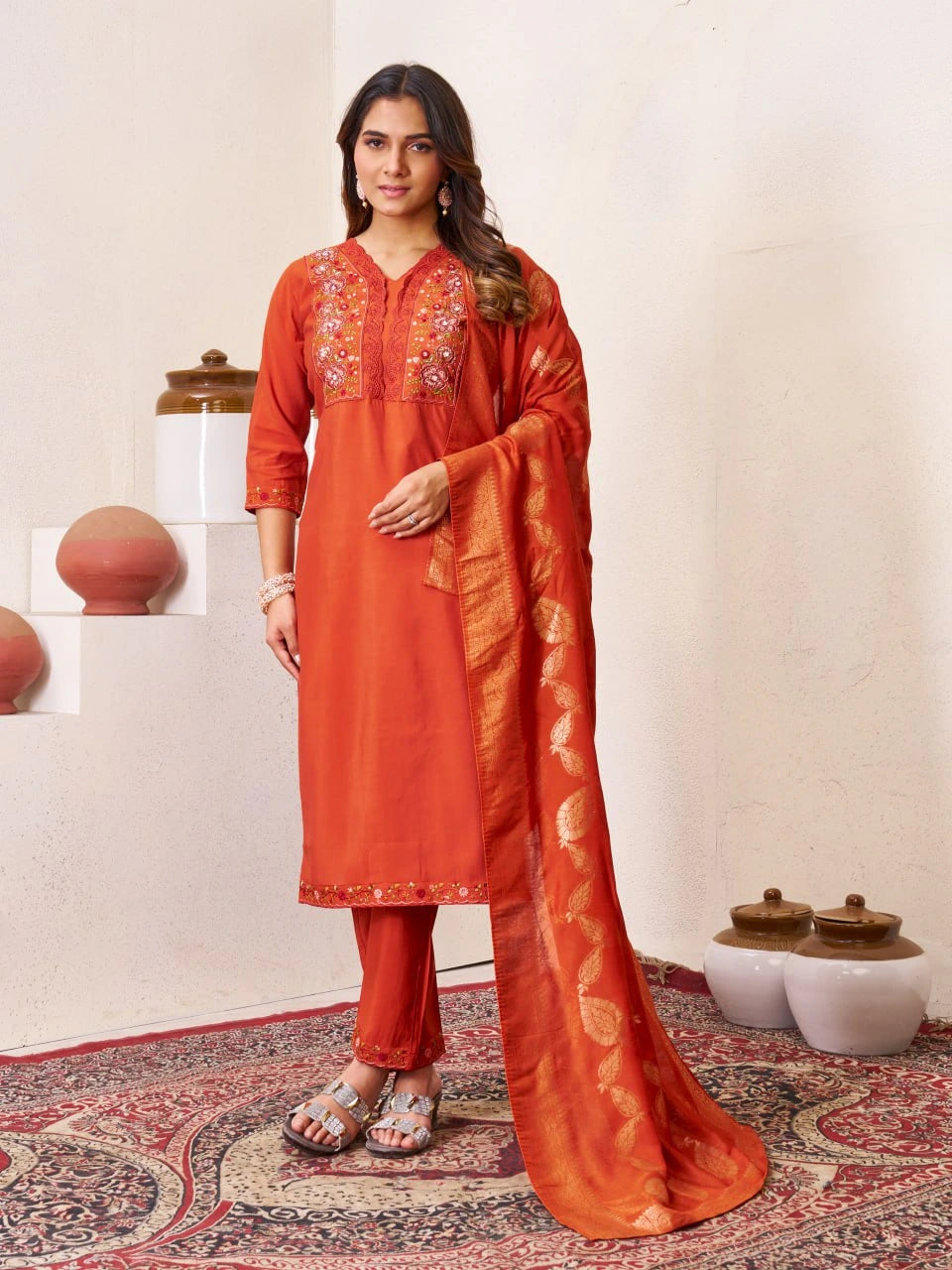 Woman wearing an orange traditional outfit with embroidery, standing on a patterned rug.